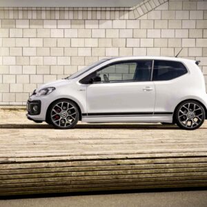 Side view of a white hatchback car parked along a light brick wall, with visible VW emblem on the grille and stylish wheels.