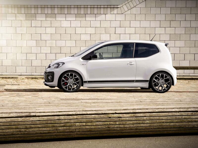 Side view of a white hatchback car parked along a light brick wall, with visible VW emblem on the grille and stylish wheels.