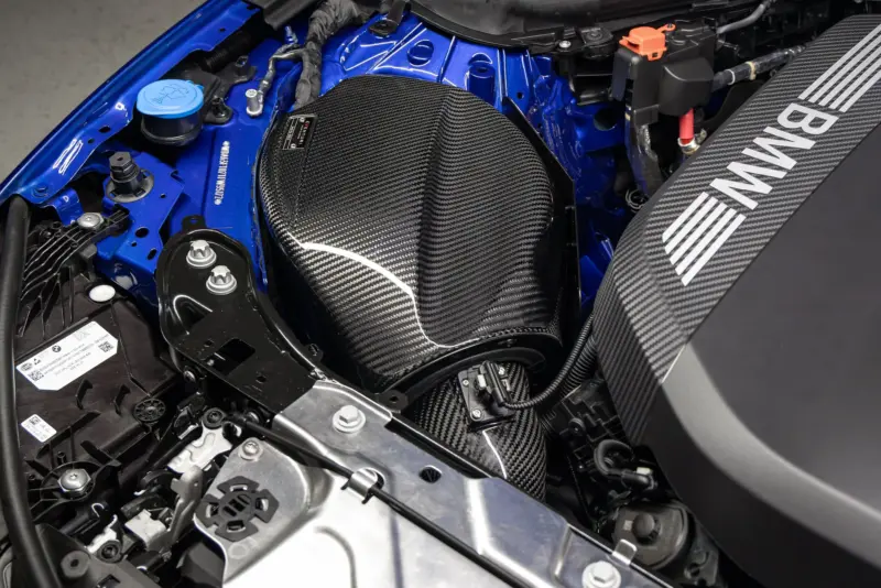 Close-up of a blue BMW engine bay featuring a carbon fiber intake cover and visible battery area.