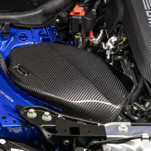 Close-up of a car engine bay with a blue body, black carbon-fiber panels, and exposed wiring; an orange connector and various hoses visible near the top.