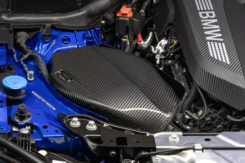 Close-up of a car engine bay with a blue body, black carbon-fiber panels, and exposed wiring; an orange connector and various hoses visible near the top.