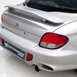 Rear view of a silver sports car with a large spoiler and red taillights, showing a dealership-style license plate area reading 'MOTOR DROME DESIGN'