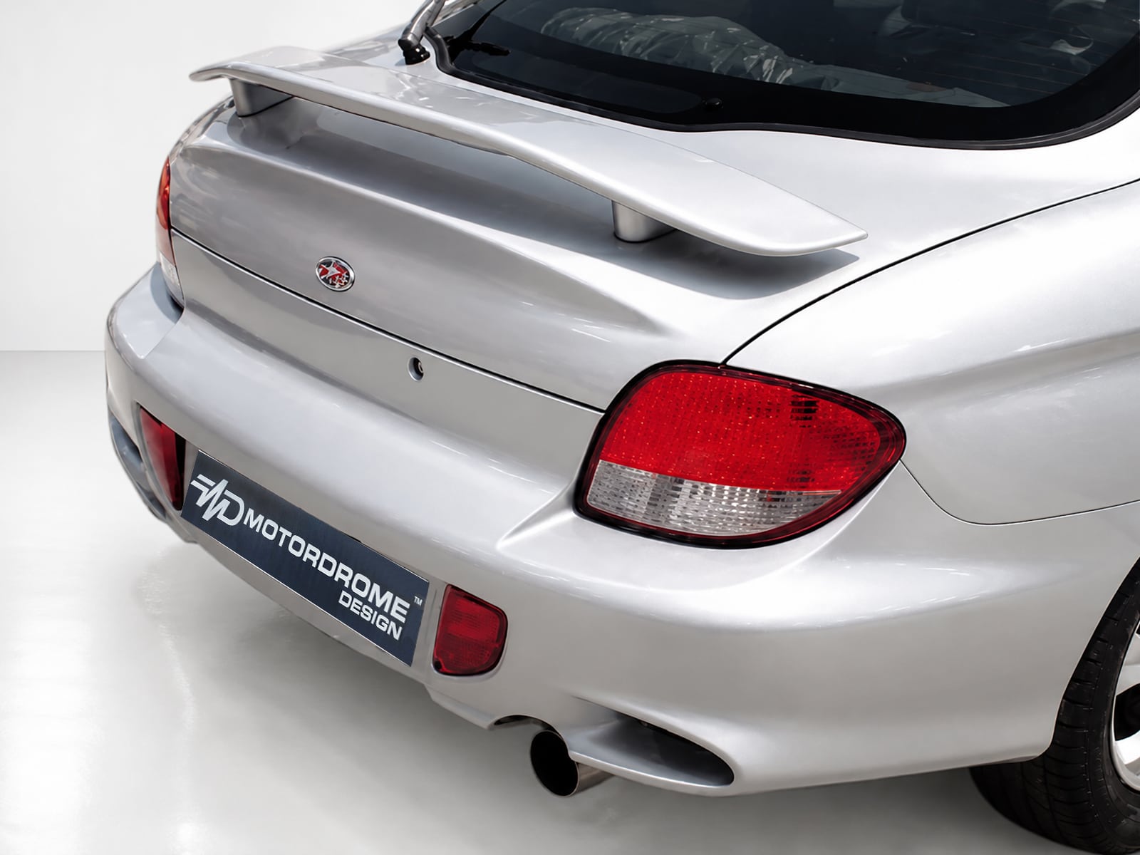 A-171_1jpg - OPSHOLDERS Rear view of a silver sports car with a large spoiler and red taillights, showing a dealership-style license plate area reading 'MOTOR DROME DESIGN'