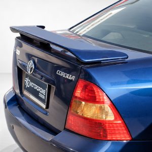 Rear view of a blue Toyota Corolla with a spoiler and prominent taillight in a showroom setting, showing the trunk and badge clearly.