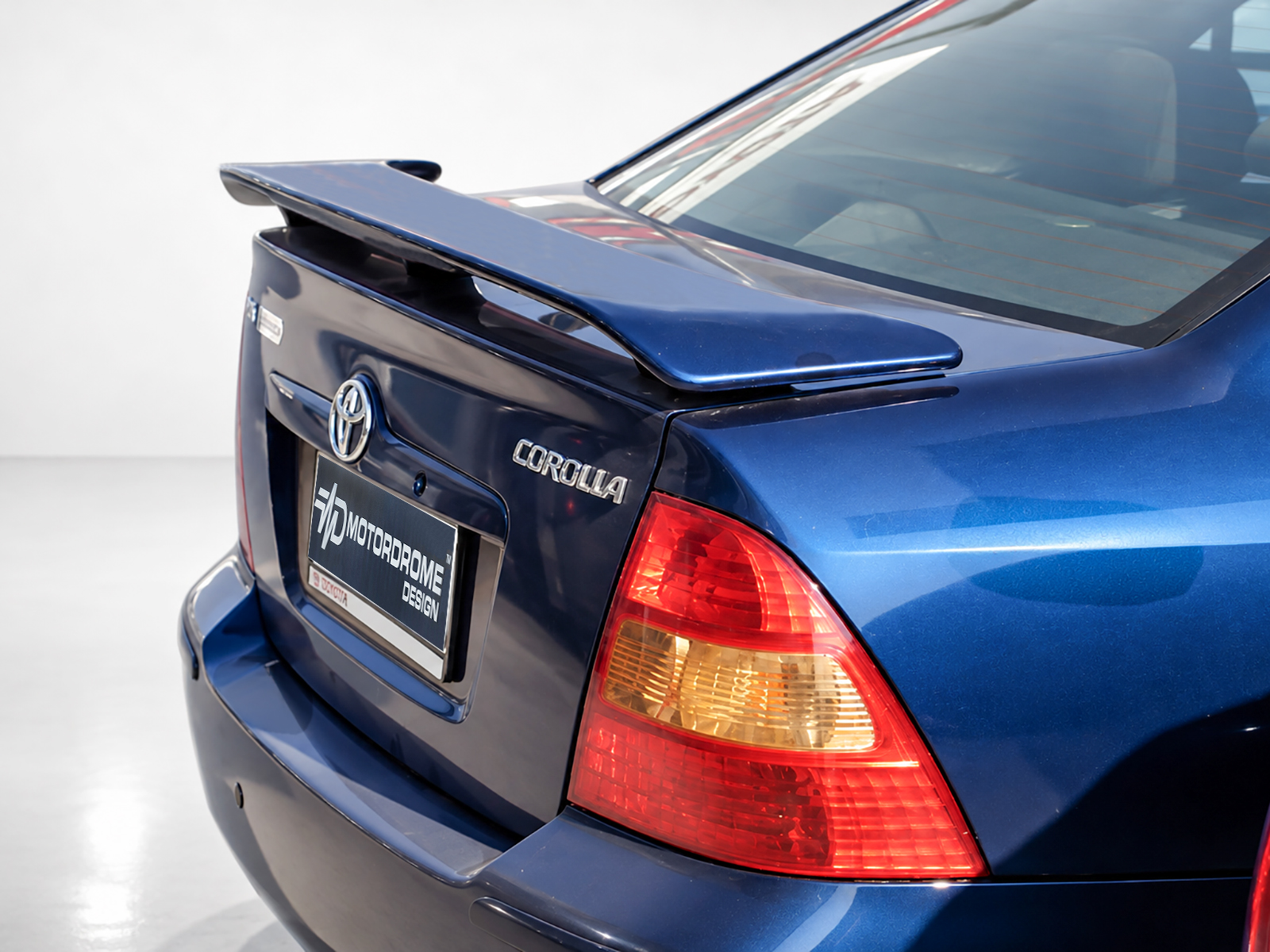 A-218_1jpg - OPSHOLDERS Rear view of a blue Toyota Corolla with a spoiler and prominent taillight in a showroom setting, showing the trunk and badge clearly.