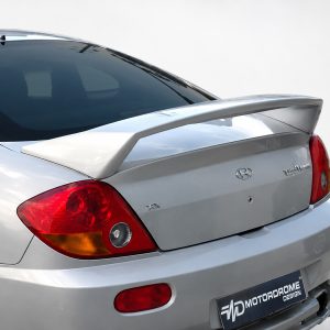 Rear of a silver Hyundai Tiburon with a large rear spoiler and red taillights on a light background.