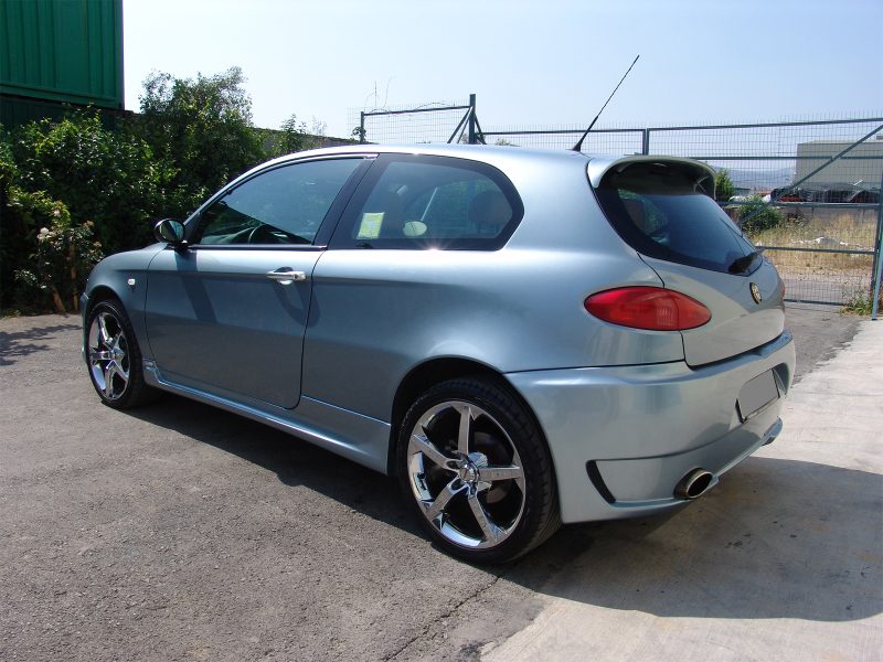 Blue hatchback car parked on a paved lot beside green shrubs, with a chain-link fence in the background.