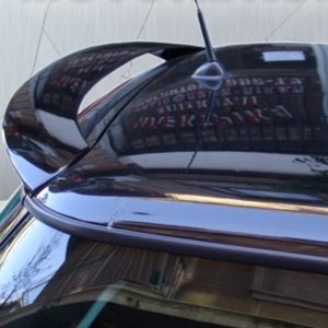Close-up of a glossy black car hood with chrome trim and antenna, reflecting the surrounding buildings and sky.