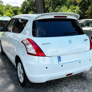 Rear view of a white Suzuki Swift hatchback parked on a gravel lot, with another car nearby and greenery in the background.