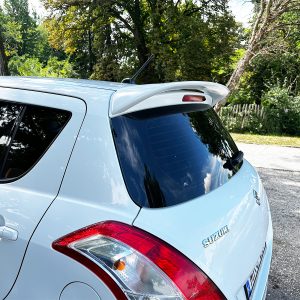 Rear three-quarter view of a light blue Suzuki hatchback with a roof spoiler, parked on a tree-lined street.