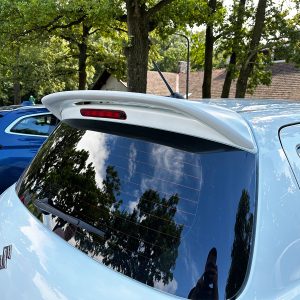 Rear view of a light-blue hatchback with a white roof spoiler and rear windshield wiper, parked among trees.