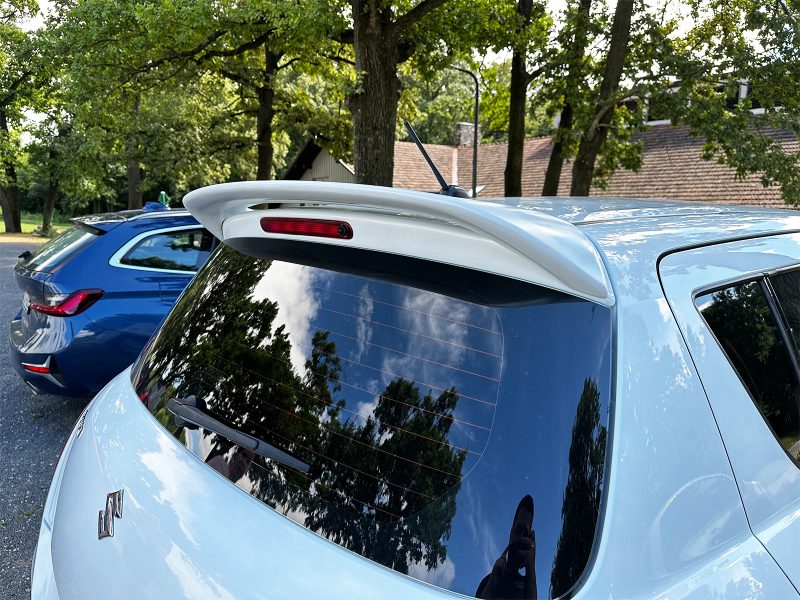 Rear view of a light-blue hatchback with a white roof spoiler and rear windshield wiper, parked among trees.