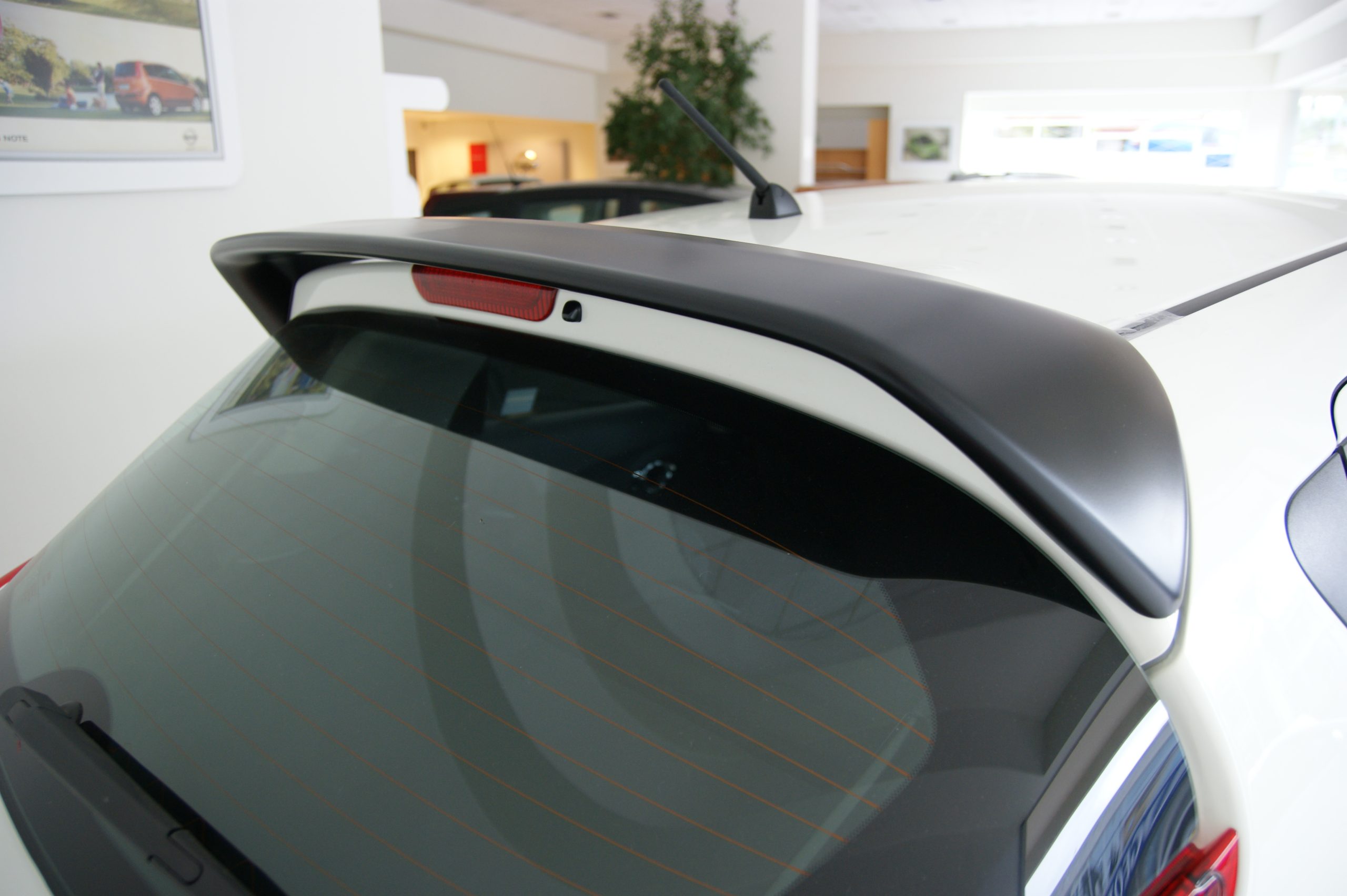 SONY DSC - OPSHOLDERS Close-up of a white car's rear window, roof spoiler, antenna, and taillight in a showroom setting