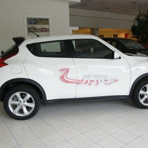 SONY DSC - OPSHOLDERS White Nissan Juke SUV on showroom floor with pink logo decal reading 'NEW NISSAN JUKE' along the side, indoors with tiled floor and other cars in background