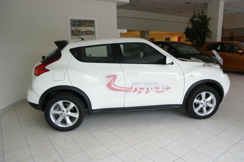 SONY DSC - OPSHOLDERS White Nissan Juke SUV on showroom floor with pink logo decal reading 'NEW NISSAN JUKE' along the side, indoors with tiled floor and other cars in background