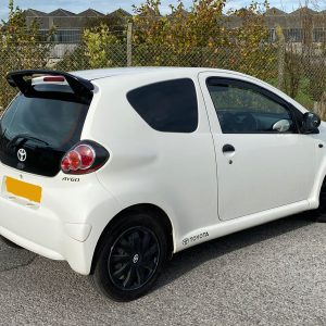 White Toyota Aygo hatchback seen from the rear-left, with a spoiler and tinted rear window parked on a street.
