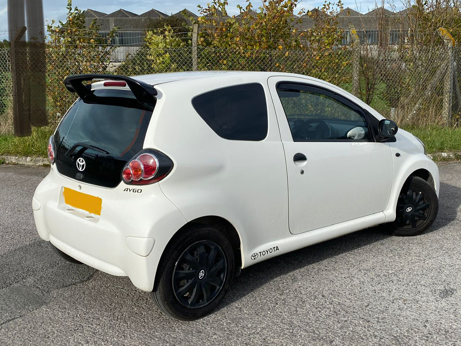 A-387_1jpg - OPSHOLDERS White Toyota Aygo hatchback seen from the rear-left, with a spoiler and tinted rear window parked on a street.