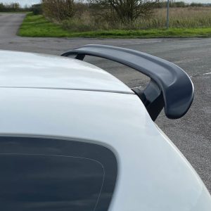 A-387_2jpg - OPSHOLDERS White car rear with a large dark gray spoiler, photographed near a rural road with grassy areas in the background.