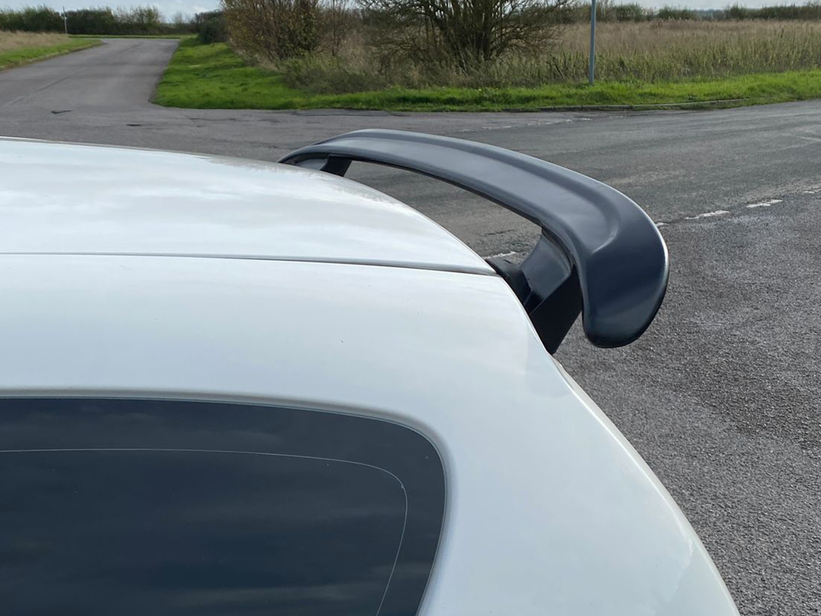 A-387_2jpg - OPSHOLDERS White car rear with a large dark gray spoiler, photographed near a rural road with grassy areas in the background.