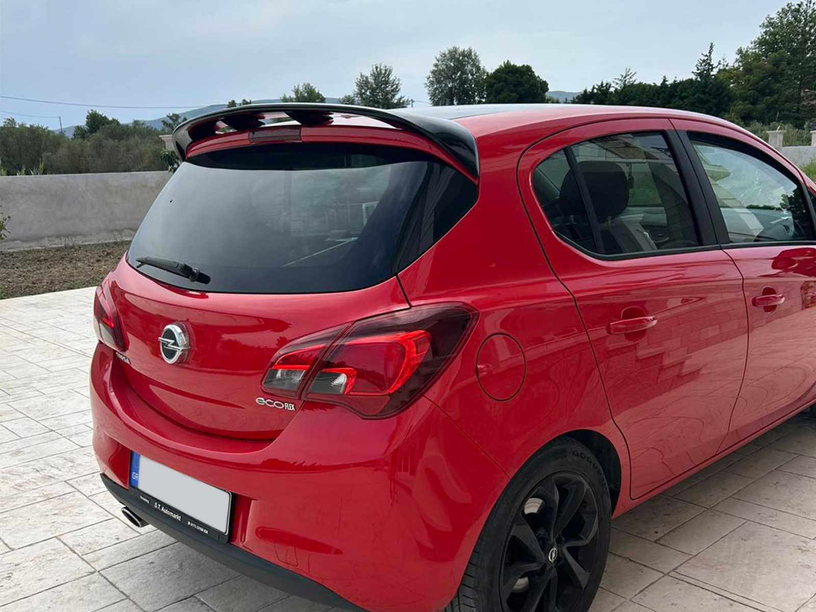 A-408_1jpg - OPSHOLDERS Red hatchback car parked on a light tiled driveway, rear view showing the Opel badge and a small rear spoiler on the roof edge.
