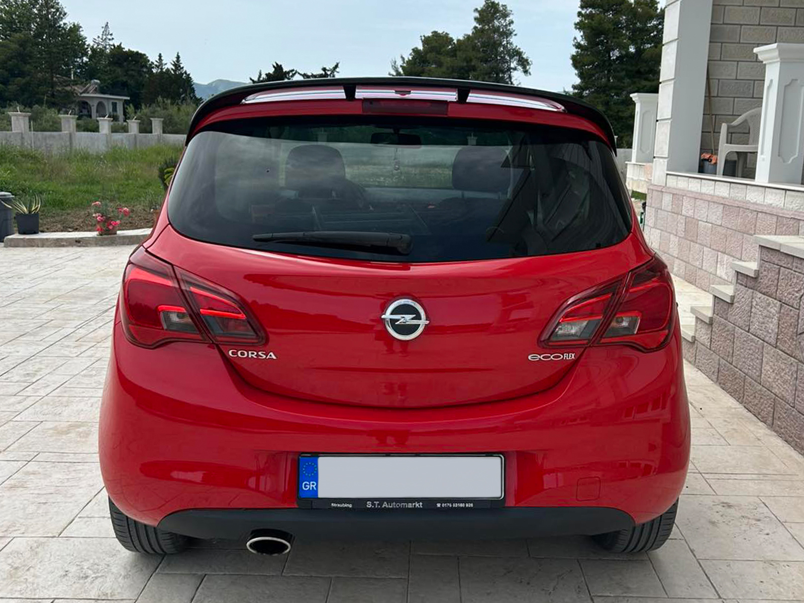 A-408_2jpg - OPSHOLDERS Rear view of a red Opel Corsa EcoFlex parked on a tiled driveway beside a house. The trunk, rear lights, and exhaust are visible with a Greek license plate.