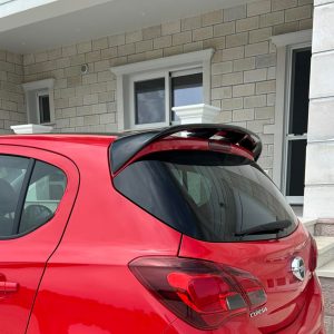 A-408_3jpg - OPSHOLDERS Rear three-quarter view of a red hatchback parked in front of a beige brick house with steps and a stone column, showing the car's spoiler and rear lights.