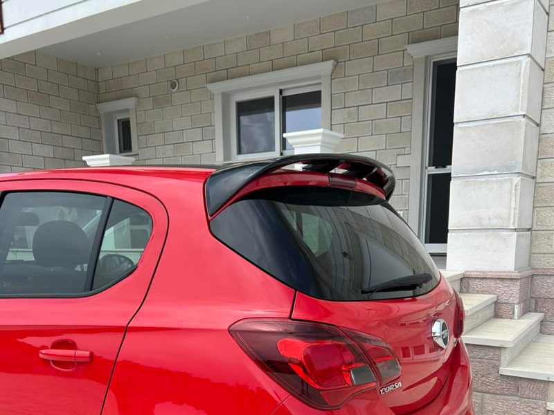 A-408_3jpg - OPSHOLDERS Rear three-quarter view of a red hatchback parked in front of a beige brick house with steps and a stone column, showing the car's spoiler and rear lights.