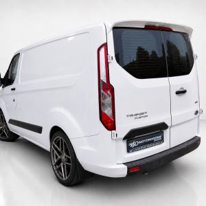 White Ford Transit Custom van photographed from a rear-left angle in a bright studio, showing black aftermarket wheels and dark rear windows.