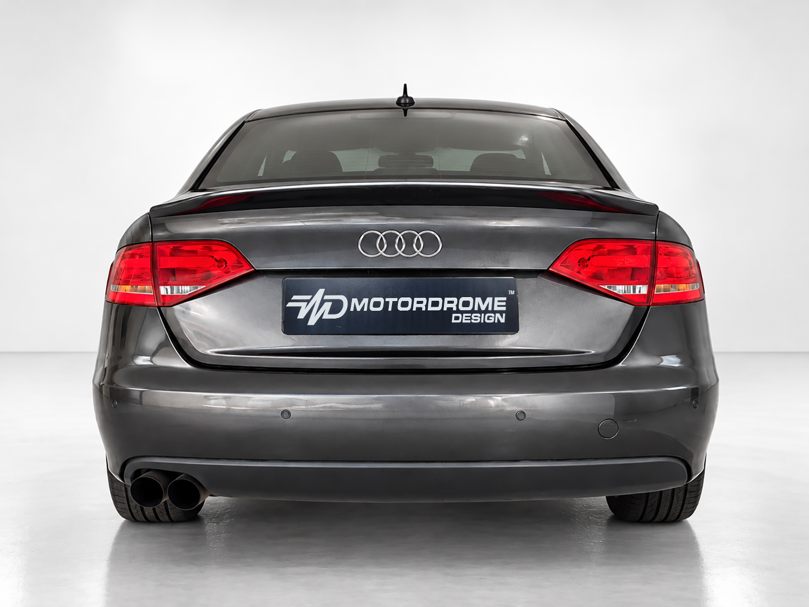 A-443_2jpg - OPSHOLDERS Rear view of a gray Audi sedan in a studio, showing taillights, dual exhausts, and a MOTORDROME DESIGN license plate.