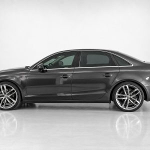 A-443_4jpg - OPSHOLDERS Side profile of a dark gray four-door sedan with large alloy wheels on a reflective studio surface, showing chrome trim.