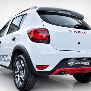A-445_3jpg - OPSHOLDERS Rear three-quarter view of a white hatchback with red DACIA badge and Motordrome Design plate, indoors.