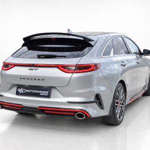 Rear three-quarter view of a silver sport sedan in a studio, showing red taillights, quad exhausts, and a PROCEED badge on the trunk.