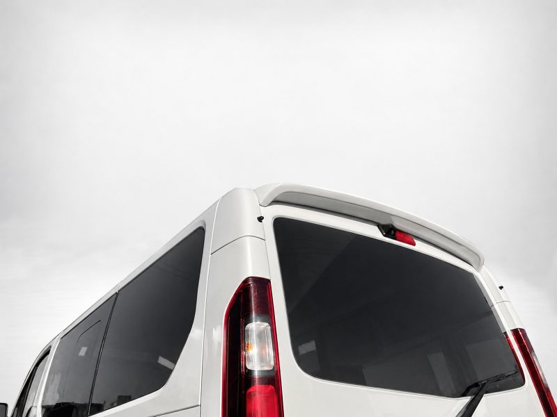 A-455_5jpg - OPSHOLDERS Rear corner of a white van with dark rear windows and vertical red taillights against a pale sky.