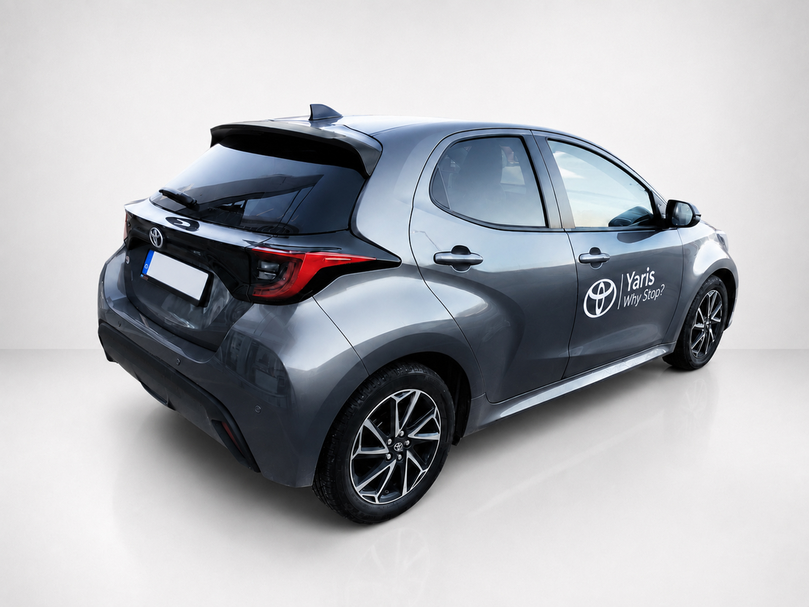 A-463_2jpg - OPSHOLDERS Rear three-quarter view of a gray Toyota Yaris hatchback with 'Yaris Why Stop?' branding on the side, in a studio setting.