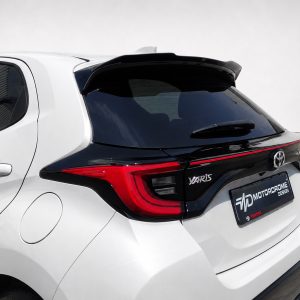 Rear view of a white Toyota Yaris hatchback with a black roof spoiler and red taillights, showing the brand emblem and a dealership plate frame.