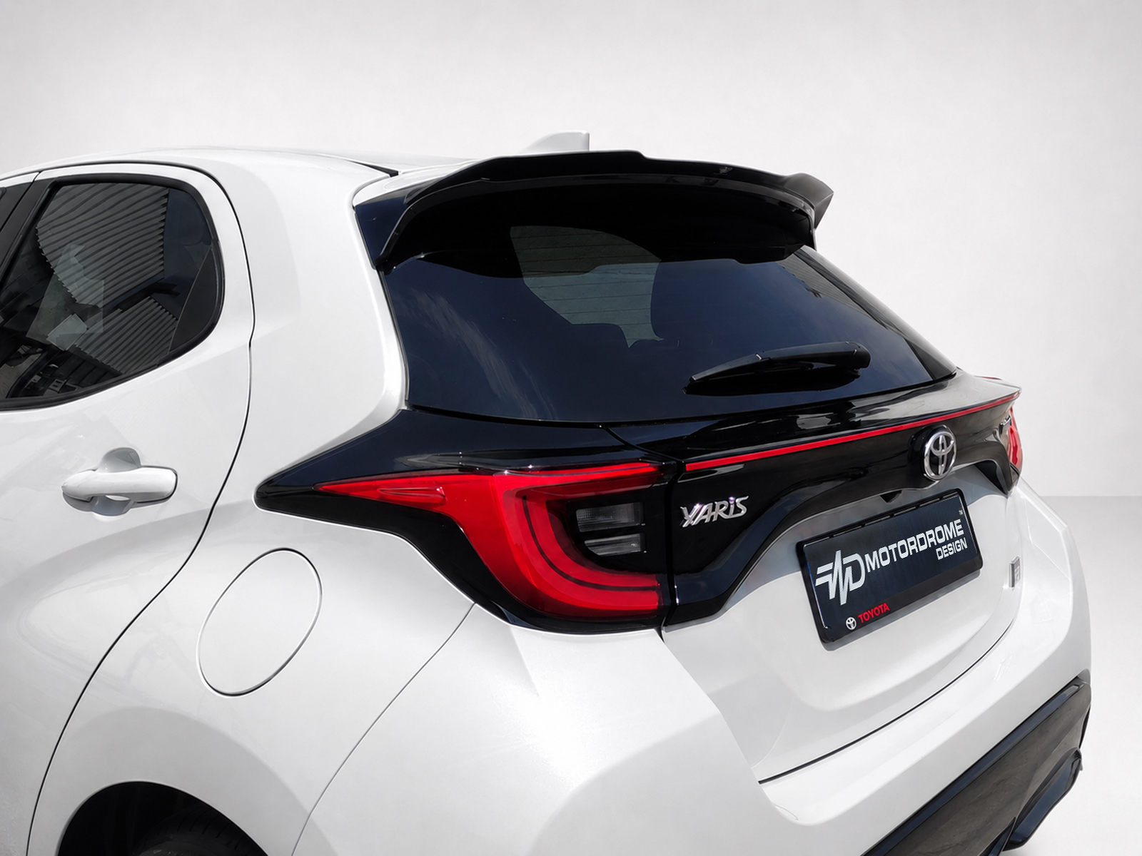 A-474_1-1jpg - OPSHOLDERS Rear view of a white Toyota Yaris hatchback with a black roof spoiler and red taillights, showing the brand emblem and a dealership plate frame.