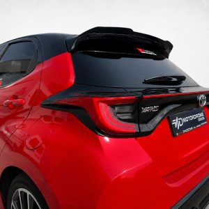 Red Toyota Yaris rear three-quarter view with black roof spoiler and glossy black rear hatch trim, showing a dealership plate reading MOTORDROME DESIGN.