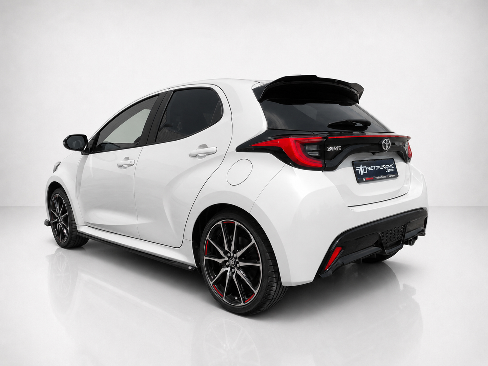 A-474_2-1jpg - OPSHOLDERS White Toyota Yaris hatchback, rear three-quarter view in a studio setting with black roof spoiler and red-accented alloy wheels.