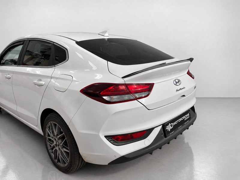 A-477_4jpg - OPSHOLDERS Rear three-quarter view of a white Hyundai sedan with a spoiler, in a studio setting with tinted rear window and alloy wheels.