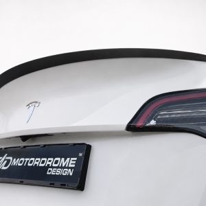 Rear view of a white car showing a black lip spoiler, sleek taillight, and a Motordrome Design license plate badge on a white backdrop.