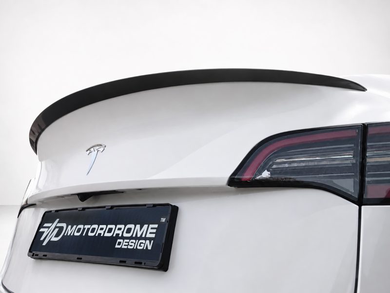 Rear view of a white car showing a black lip spoiler, sleek taillight, and a Motordrome Design license plate badge on a white backdrop.