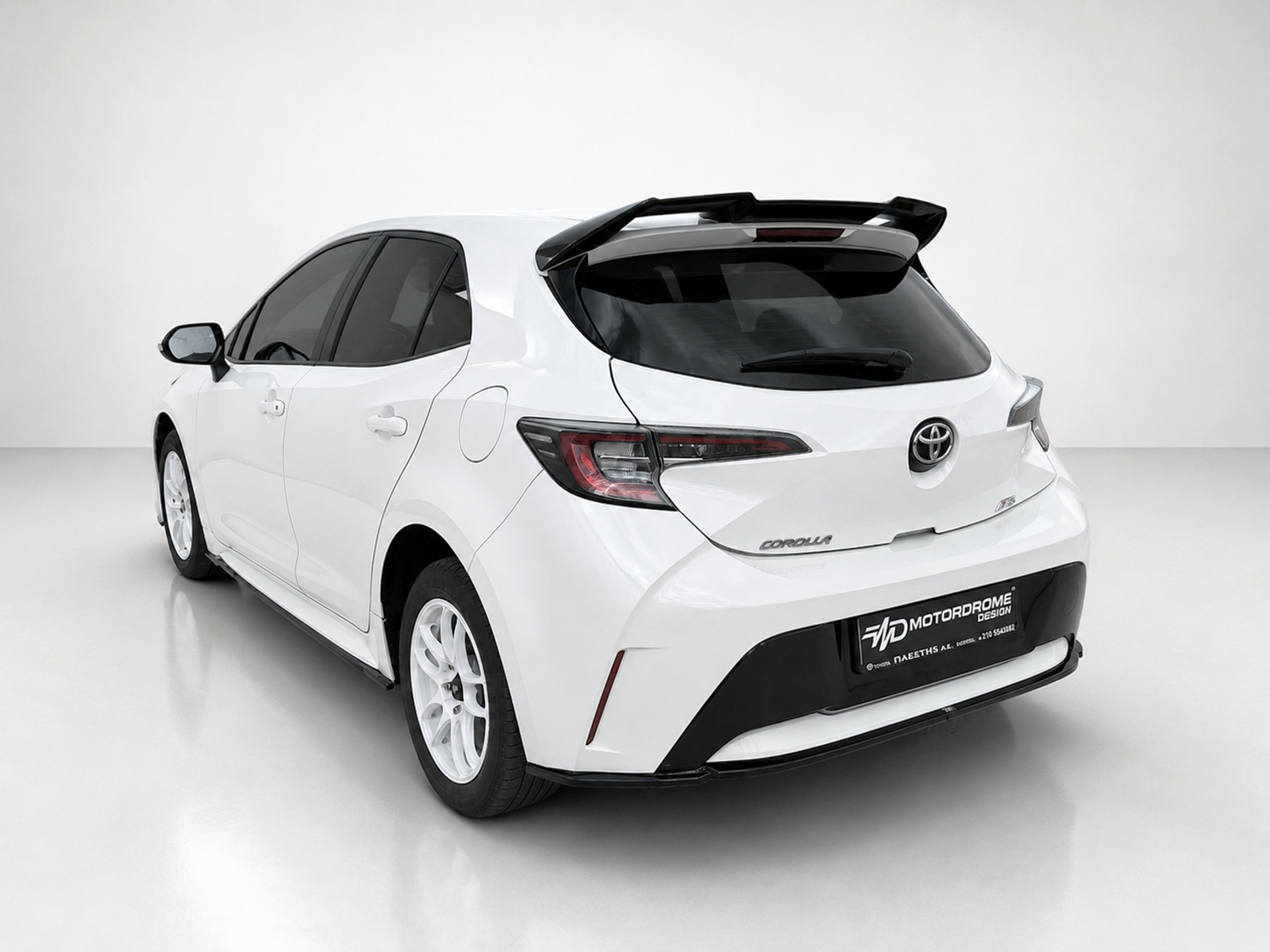 A-511_2jpg - OPSHOLDERS Rear three-quarter view of a white Toyota Corolla hatchback in a studio setting, front wheels turned slightly left with a spoilered roofline visible.