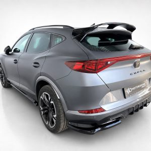 Silver CUPRA SUV shown from a rear‑left three‑quarter angle in a studio, with a prominent rear wing and red taillights visible.