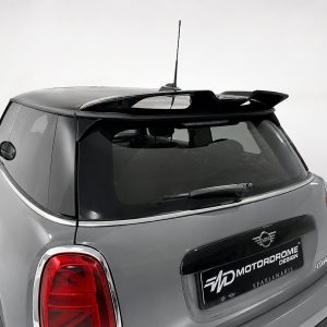 Grey Mini Cooper rear view with roof spoiler, antenna, and rear windshield wiper in a studio setting outside the vehicle brand plate shows 'Motoradrome Design' (advertising image).