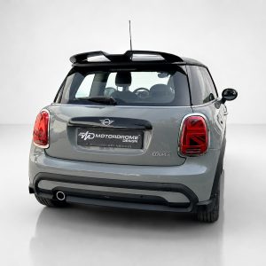 Rear view of a gray Mini Cooper with a custom Motordrome Design plate and red tail lights in a studio setting? and a single exhaust visible on the left.