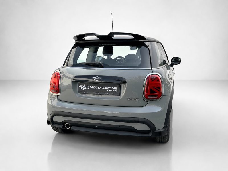 Rear view of a gray Mini Cooper with a custom Motordrome Design plate and red tail lights in a studio setting? and a single exhaust visible on the left.