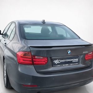 Rear view of a dark gray BMW 316i sedan, showing red taillights and a dealership license plate in a bright studio setting.