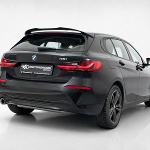 Rear three-quarter view of a dark gray BMW 116i hatchback in a studio setting with a rear spoiler visible on top of the tailgate.