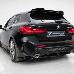 Rear three-quarter view of a black BMW M135i xDrive hatchback in a studio setting with red taillights and aftermarket rear bumper.