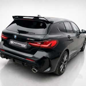 Rear three-quarter view of a black BMW M135i with large spoiler and aggressive rear diffuser in a studio setting, showing red taillights and dual exhausts.